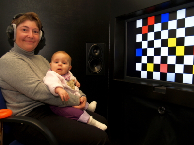 Baby on the mother's lap. The mother is wearing headphones. In the background there is a loudspeaker and a large monitor with a colourful pattern on it.